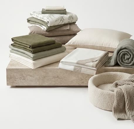 Neutral sage and beige folded bedding sets with pillows and knit throw on stone plinth